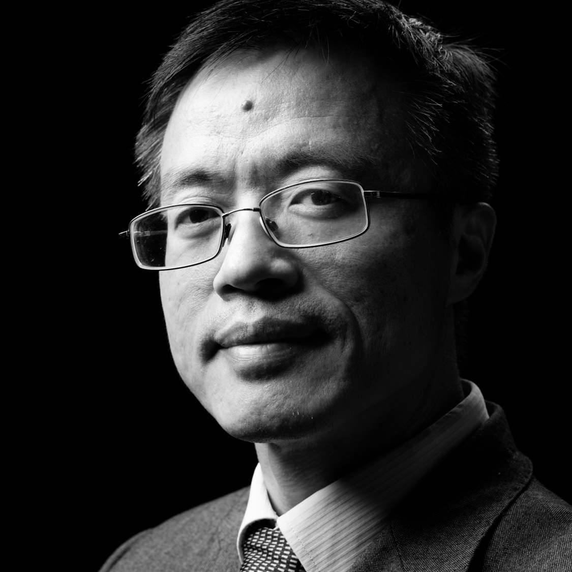 A monochrome image of a man with dark hair and glasses wearing a suit that is looking at the camera.